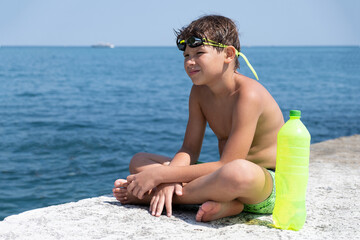 A cute boy in swimming glasses and shorts with a bottle of water sits on a pier on the seashore.