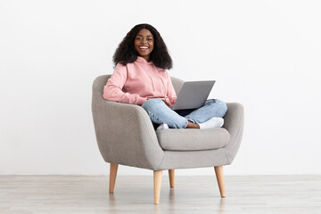 Naklejka premium Pretty black lady sitting in armchair with laptop