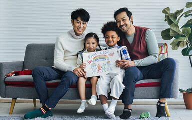 Male gay taking care adopted children who are happy diverse little Caucasian girl and African boy, happily smiling, drawing family picture, sitting on sofa in living room at home. LGBT, kids Concept.