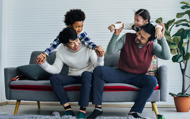 Male gay taking care adopted children who are happy diverse little Caucasian girl and African boy, hugging, playing toys together with fun, sitting on sofa in living room at home. LGBT, kids Concept.