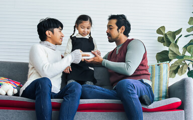 Asian male Gay couple taking care adopted little daughter girl, feeding her breakfast in the morning, smiling with happiness, sitting on sofa in living room at home. LGBT, Family and Kid concept.