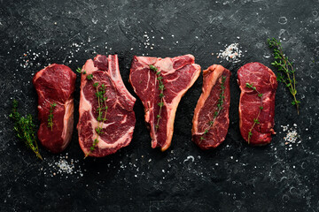 A variety of raw beef steaks for grilling with seasonings: T-bone, striploin steak, ribeye. Top view. On a black stone background.