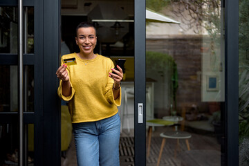 Portrait of smiling woman using phone for online payment on doorstep of her home shopping online from home leaning on door frame using credit card looking at camera cheerful and happy