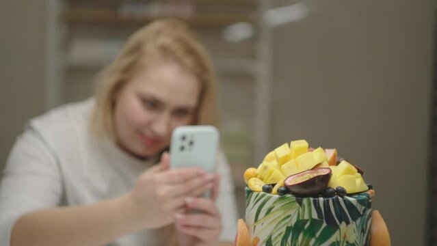 Woman confectioner taking photo of rotating cake decorated with mango, passion fruit, grapefruit and blueberry. Bakery process in slow motion.