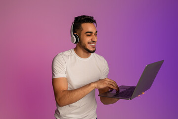 Positive millennial Arab man in wireless headphones using laptop pc for online work or...