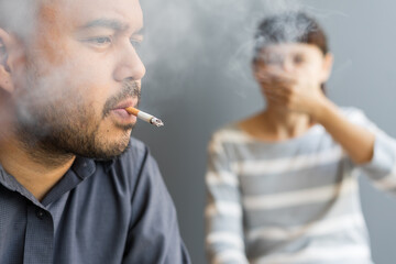 Husband smoking cigarette and wife choking of smoke. Man smoking cigarette and woman is covering her face and a lot of smoke around smelling pollution. Passive stop smoking cigarettes campaign.