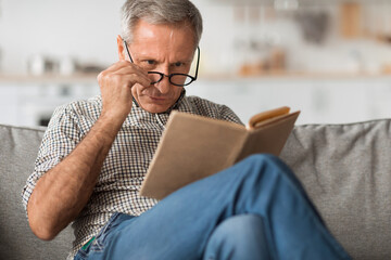 Senior Man With Poor Eyesight Reading Book Above Eyeglasses Indoor - obrazy, fototapety, plakaty