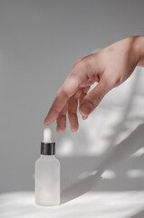 The hand touches a cosmetic bottle with essential oil or serum. White cosmetic bottle and female hand on a white background with shadows