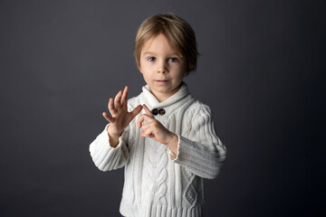 Fototapeta premium Cute little toddler boy, showing gesture in sign language on gray background, isolated image, child showing hand sings
