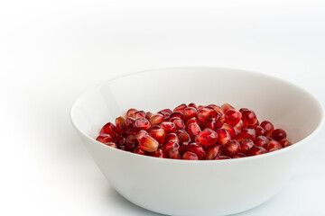 Red pomegranate seeds in a white plate, on the white background. Fresh juicy pomegranate seeds isolated on white. Close-up, side view, selective focus, copy space