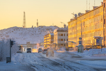 Street in a northern city in the Arctic. Winter city landscape. Cars drive along a snow-covered street. Buildings at sunset. Telecommunication towers on the mountain. Anadyr, Chukotka, Siberia, Russia