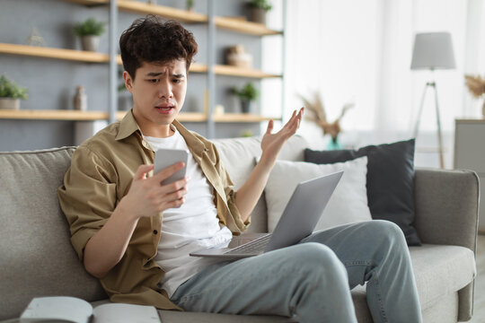 Confused asian man looking at mobile phone using laptop