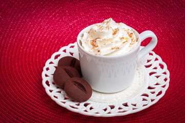 White mug with whipped cream and chocolate cookies on a white plate and a red background