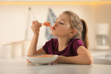 Cute little girl eating tasty pasta at table in kitchen