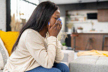 Upset african-american woman sitting on the sofa in grief, closing eyes with palms and crying, disappointed female feels despair, worrying, depression, loneliness, suffering from strong headache