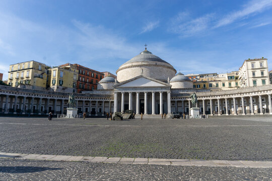 Piazza del Plebiscito in Naples, with the blue sky and some high clouds, with the colored houses behind. View of the basilica in the foreground.