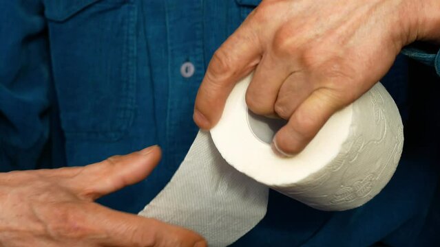 The hands of a pensioner hold a roll of toilet paper. An elderly man unwinds and tears off toilet paper. Digestive problems, constipation and diarrhea in the elderly
