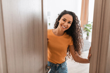 Cheerful young curly lady receiving guests at front door - Powered by Adobe