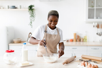 Glad happy young african american female in apron make dough for baking, prepares eat in...