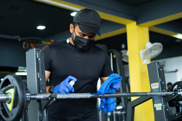 Young man with medical face mask and hand gloves sanitizing or wiping sweat of gym equipments due to coronavirus - concept of covid-19 hygiene safety measures, protection from infection at fitness