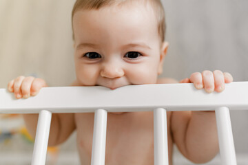 A cute baby is standing in the crib and biting the railing. Teeth eruption concept. Cute toddler...
