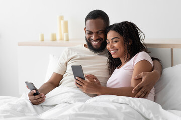 Smiling happy young african american guy hugs lady and typing, surfing, chatting on smartphones on comfortable bed