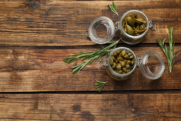 Fototapeta na wymiar Tasty capers in jars and rosemary on wooden table, flat lay. Space for text