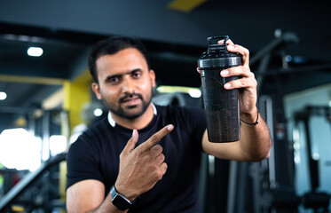 focus on bottle, Confident indian bodybuilder shacking protein bottle and showing by looking at...