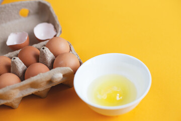 raw eggs in a package next to white bowl with one broken egg in it on a yellow background