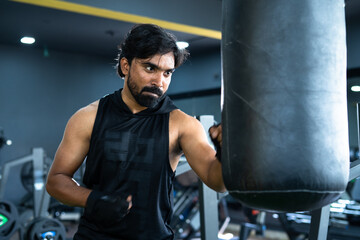 Angry young Indian athlete punching boxing bag at gym - concept of motivational, hardworking,...