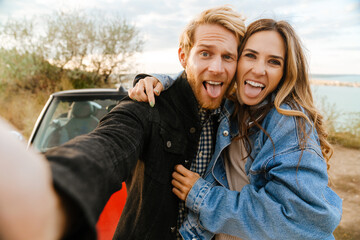 White couple making fun while taking selfie photo by car during trip