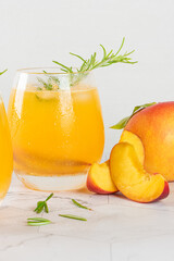 Healthy summer juice - peach cocktail with ice, rosemary twig, sugar rim, fruit slices in misted glass in elegant white interior on white marble countertop. Closeup.