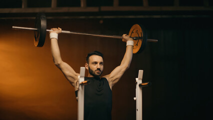 Fototapeta na wymiar Muscular sportsman training with barbell near stand on dark background