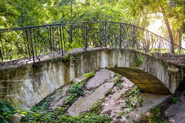 Fototapeta premium Old concrete arched footbridge with wrought iron railings against backdrop of green foliage. Dry river with concrete slabs. Ancient park in summer.