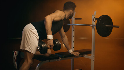 Athletic sportsman training with dumbbell near barbell on dark background
