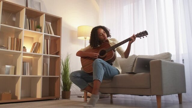 Song learning. Guitar playing. Music hobby. Focused woman musician practicing chords enjoying string acoustic instrument leisure on couch at living room at home free space.