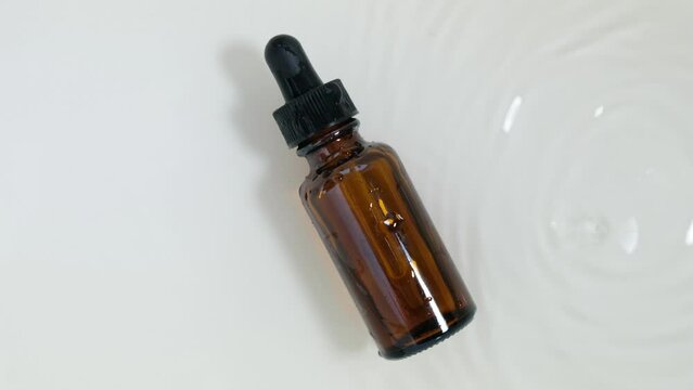 Glass cosmetic bottle with pipette lies on the surface of the water. Top view of drop falls into water and diverging circles of water on white background. Sample of packaging for design, advertising. 
