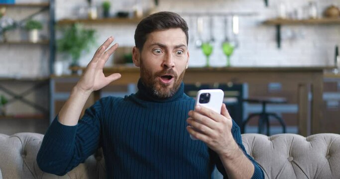 Excited and pleasantly surprised handsome man rejoices at the message on his smartphone. Young male looking at mobile phone and feel amazed happy and saying WOW on home background.
