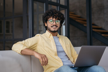Handsome confident Indian programmer using laptop computer looking at camera working from home....