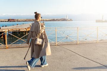 Young adult trendy stylish beautiful caucasian happy smiling woman enjoy walking by Yalta sea embankment on warm sunny day. Female person portrait wear jeans biege trench coat on urban city street