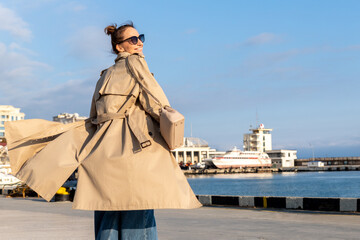 Young adult trendy stylish beautiful caucasian happy smiling woman enjoy walking by Yalta sea embankment on warm sunny day. Female person portrait wear jeans biege trench coat on urban city street