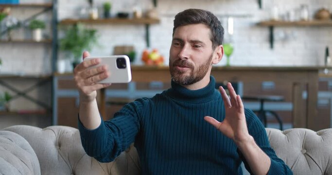 Young handsome bearded man holding smartphone in outstretched arm, making conference over video call, smiling greeting his interlocutor, sitting on sofa at home. Online communication concept