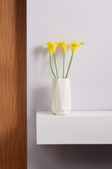 Subject shot of a graceful bouquet of tender yellow callas in a white ceramic vase. The vase with a bunch of flowers is located on the white table on the white background.