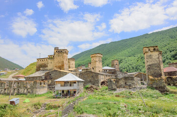 Fototapeta na wymiar View of Ushguli fortified village in Svanetia, Georgia. Grey, brown and biege medieval stone towers and old houses, green and yellow dry grass, green mountains and blue sky with clouds