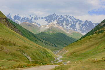 Fototapeta premium Mountain valley near Ushguli medieval fortified village in Svanetia, Georgia. Green and grassy mountain slopes, grey rocky cliffs, snow covered summits, sky with clouds, winding road