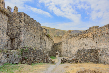 Inner yard of the Khertvisi fortress in Georgia, Caucasus. Ancient weathered medieval grey and biege stone walls and towers with windows and arch doorway, blue sky with clouds, yellow and green grass