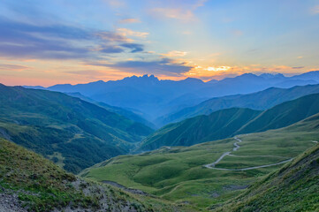 Naklejka premium View of the summer sunset above the caucasian mountains from the Bear Cross mountain pass in Khevsureti, Georgia. Blue and orange sky with clouds, green and blue haze maountains, road to Shatili