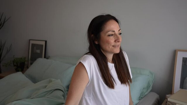 Portrait of relaxed and pensive brunette woman, sitting on bedroom bed, looking out the window and admiring the splendid day, smiling spending the weekend at home.