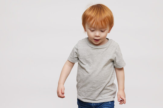 Cheerful redheaded little boy and child with beautiful ginger red hair and a grey t-shirt, looking cute smiling and being happy while he is walking and looking down. On white background
