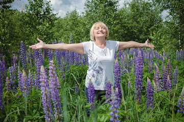 Happy attractive senior woman enjoys a walk among purple lupines in blooming field. Health, beauty...